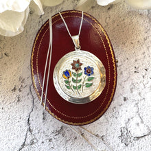 Charger l'image dans la galerie, May include: A silver locket necklace featuring a floral design in red, blue, and yellow enamel. The round locket rests on a red leather jewellery box. The chain is silver.
