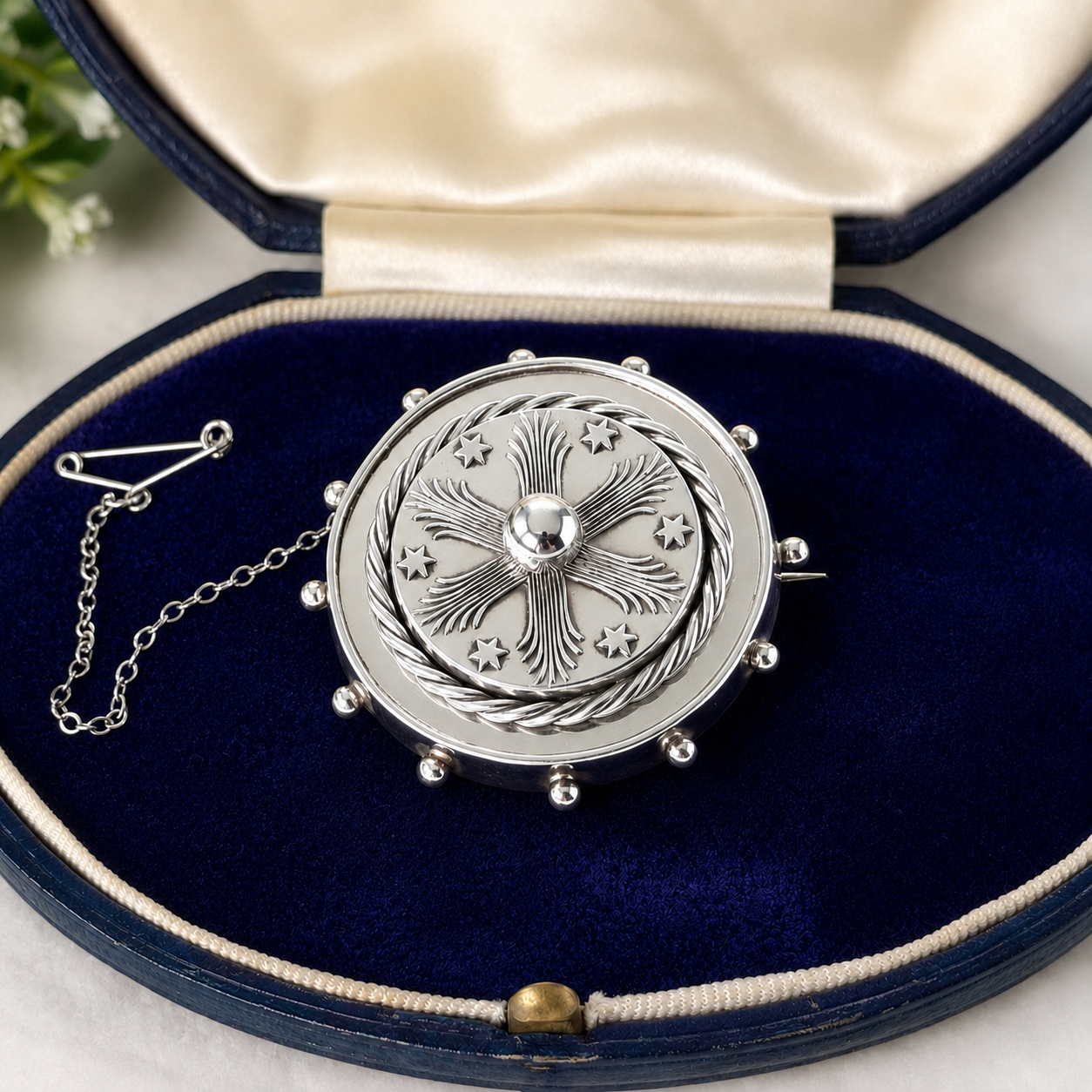 A silver-toned circular brooch with an engraved floral and star pattern, displayed inside an open navy blue velvet jewelry box with a cream satin lining.