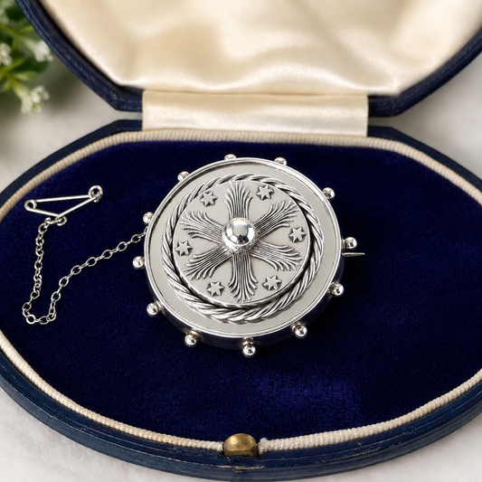 A silver-toned circular brooch with an engraved floral and star pattern, displayed inside an open navy blue velvet jewelry box with a cream satin lining.