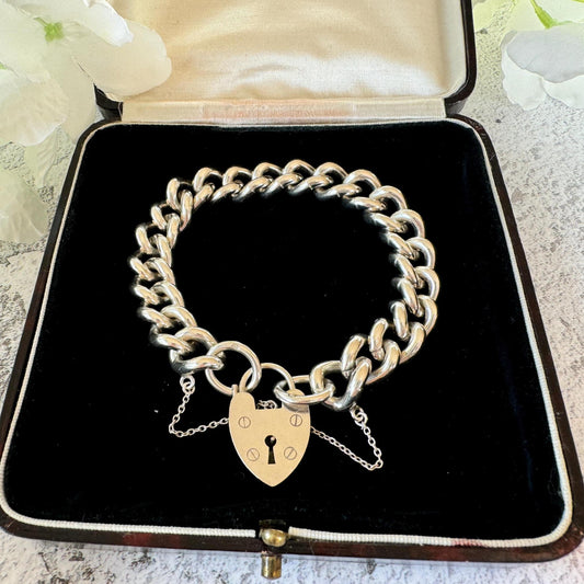May include: A silver charm bracelet featuring a heart-shaped padlock charm. The bracelet is made of thick, chunky links and is presented in a black velvet box.
